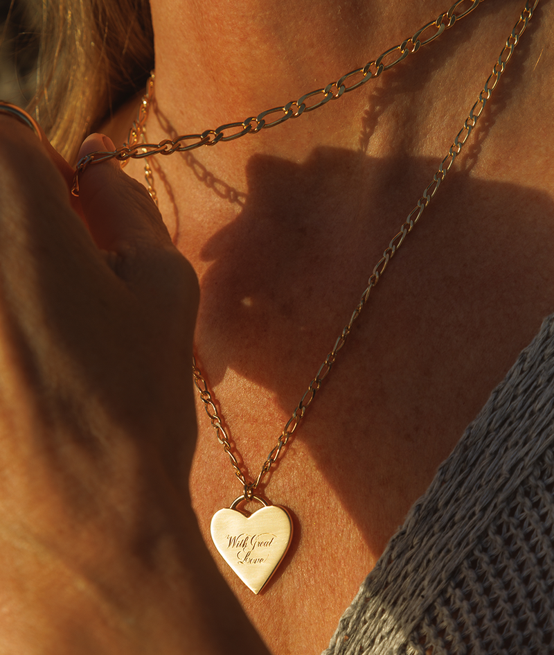 With Great Love Solid Gold Heart Pendant, with hand engraving and matte finish, worn on body on figaro chain.